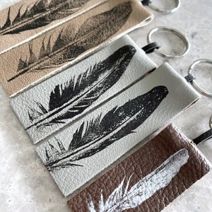 May include: Five leather keychains with different feather designs. The keychains are in various shades of brown and gray. The feathers are printed in black ink.