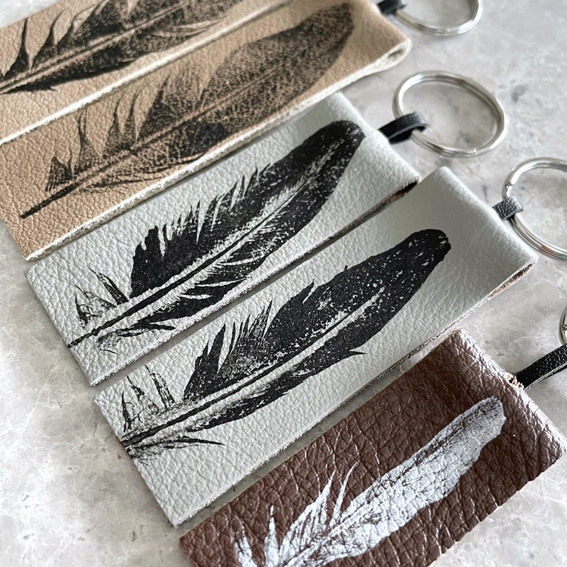 May include: Five leather keychains with different feather designs. The keychains are in various shades of brown and gray. The feathers are printed in black ink.