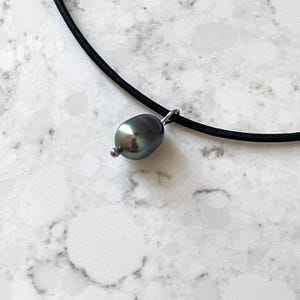 Puede incluir: Un collar de cord&oacute;n de cuero negro con un colgante de perla gris oscuro.