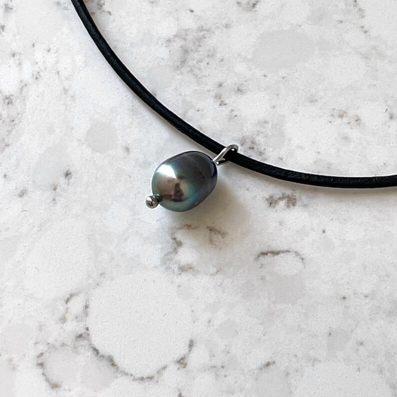 Puede incluir: Un collar de cord&oacute;n de cuero negro con un colgante de perla gris oscuro.