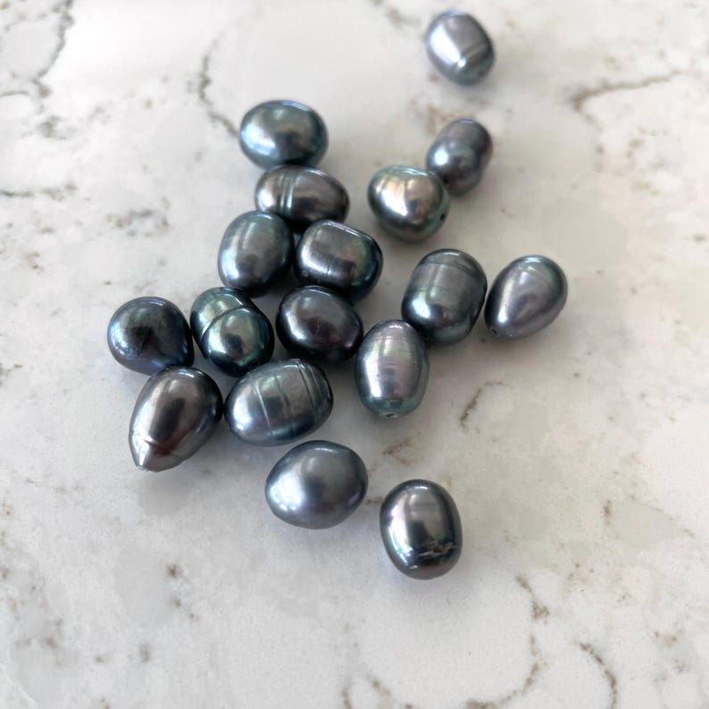 Puede incluir: Una colecci&oacute;n de perlas ovaladas de color gris oscuro, esparcidas sobre una superficie de m&aacute;rmol blanco.