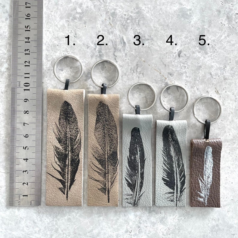 May include: Five leather keychains with a feather design. The keychains are in different colors, including brown, gray, and beige. The keychains are numbered 1 through 5.