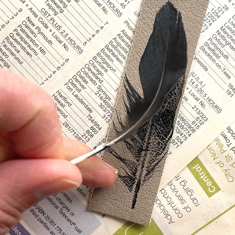 May include: A black feather with a white quill is being held by a hand over a piece of paper with a printed feather design. The paper has a list of cities and their area codes.