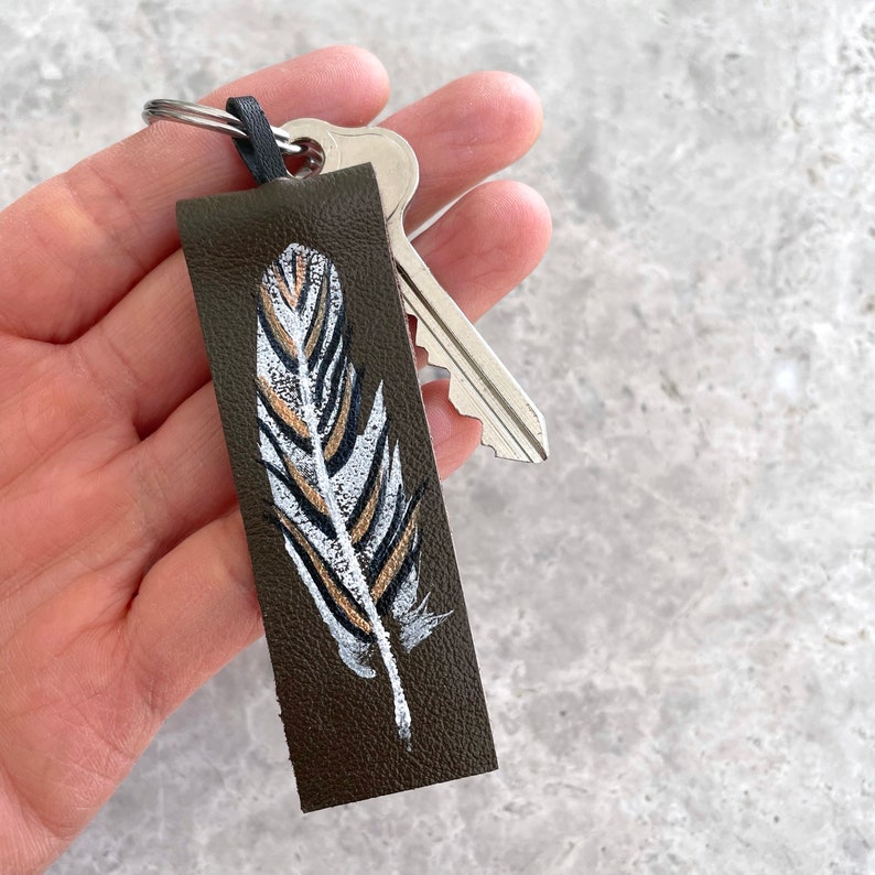 May include: A brown leather keychain with a painted white and gold feather design.