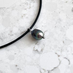 Puede incluir: Un collar gargantilla de cuero negro con un colgante de perla gris oscuro.