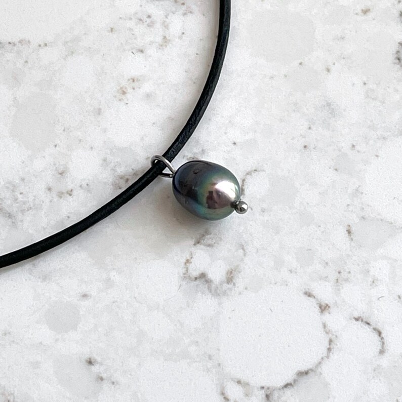 Puede incluir: Un collar gargantilla de cuero negro con un colgante de perla gris oscuro.