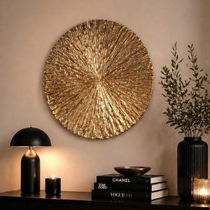 May include: A large circular gold wall art piece with a textured sunburst pattern hangs above a black sideboard. The sideboard holds a black dome lamp, a stack of books labelled Chanel and Vogue, a black vase with greenery, and a small candle.