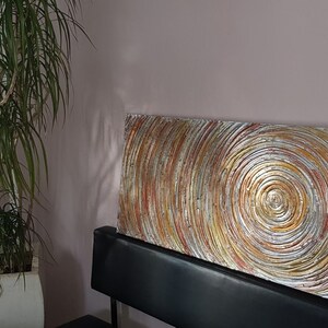 May include: Abstract wall art featuring concentric circles in shades of gold, silver, and red. The textured artwork is rectangular and rests on a black surface. A tall green plant in a white pot is to the left.