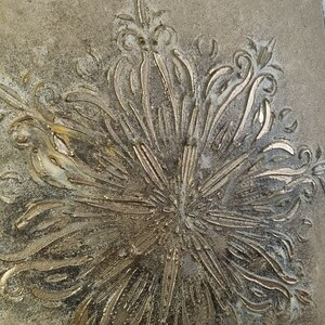 Op de afbeelding: Een goudkleurige wanddecoratie met een reli&euml;f, gegraveerd ontwerp van een gestileerde bloem. De achtergrond is lichtbruin.
