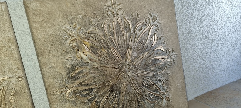 Op de afbeelding: Een goudkleurige wanddecoratie met een reli&euml;f, gegraveerd ontwerp van een gestileerde bloem. De achtergrond is lichtbruin.