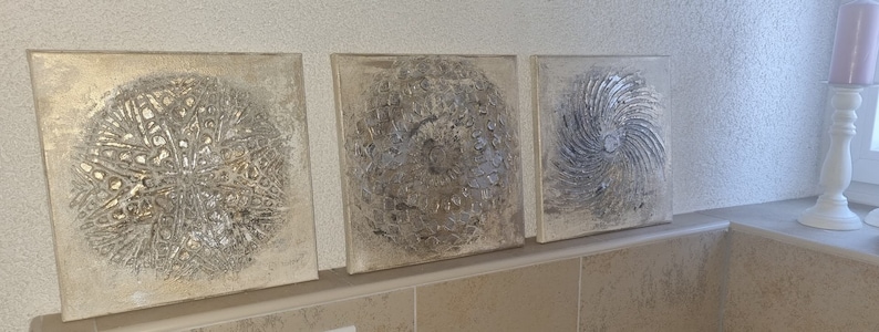 May include: Three square canvas wall art pieces with a silver and gold metallic finish. Each piece features a circular design with a textured, abstract pattern.