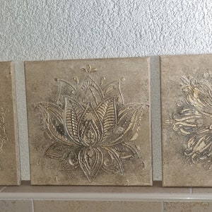 Op de afbeelding: Drie vierkante canvas wandkunststukken met een beige achtergrond en verhoogde, ge&euml;mbosseerde ontwerpen. De ontwerpen zijn ingewikkeld en bevatten een mandala, een lotusbloem en een bloemenpatroon.