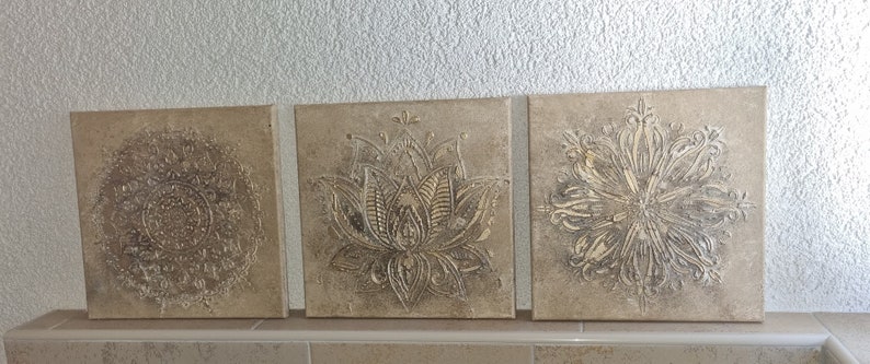Op de afbeelding: Drie vierkante canvas wandkunststukken met een beige achtergrond en verhoogde, ge&euml;mbosseerde ontwerpen. De ontwerpen zijn ingewikkeld en bevatten een mandala, een lotusbloem en een bloemenpatroon.