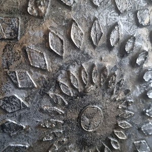 May include: A close-up of a textured, metallic wall art piece with a silver and gold color scheme. The art features a variety of geometric shapes and patterns.