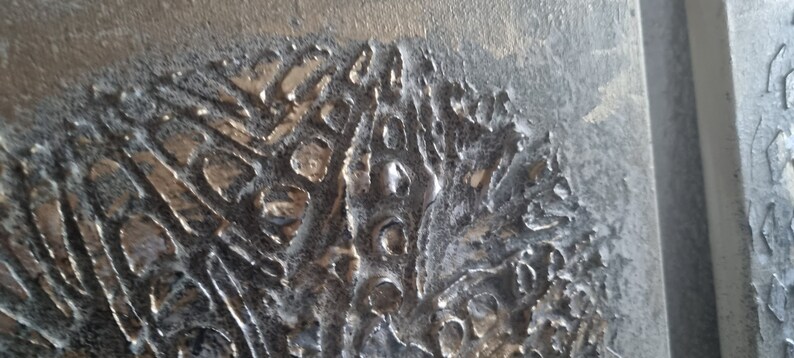 May include: Close-up of a textured, metallic surface with a silver and gray color scheme. The surface has a raised, abstract pattern that resembles a natural landscape.