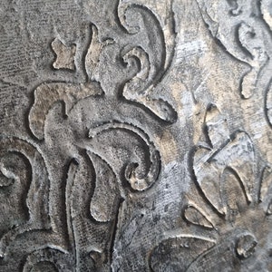 May include: A close-up of a textured, gray surface with a gold metallic design. The design features intricate swirls and flourishes.
