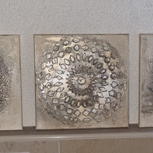May include: Three square canvas wall art pieces with a silver and gray color scheme. The center piece features a circular design with a raised texture. The left and right pieces feature a swirling design with a raised texture.