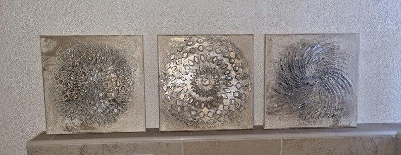 May include: Three square canvas wall art pieces with a silver and gray color scheme. The center piece features a circular design with a raised texture. The left and right pieces feature a swirling design with a raised texture.