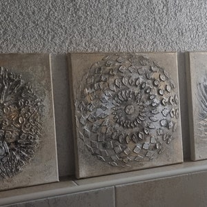 May include: Three square canvas paintings with a textured, metallic silver and gold design. Each painting features a different circular pattern.