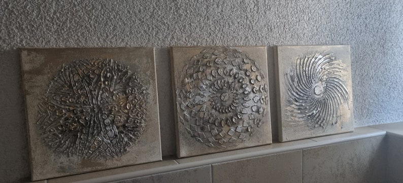 May include: Three square canvas paintings with a textured, metallic silver and gold design. Each painting features a different circular pattern.