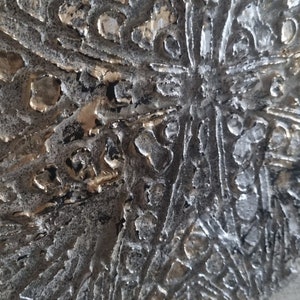 May include: A close-up of a textured, gray canvas with a raised, silver and gold design. The design is abstract and resembles a stylized flower or snowflake.