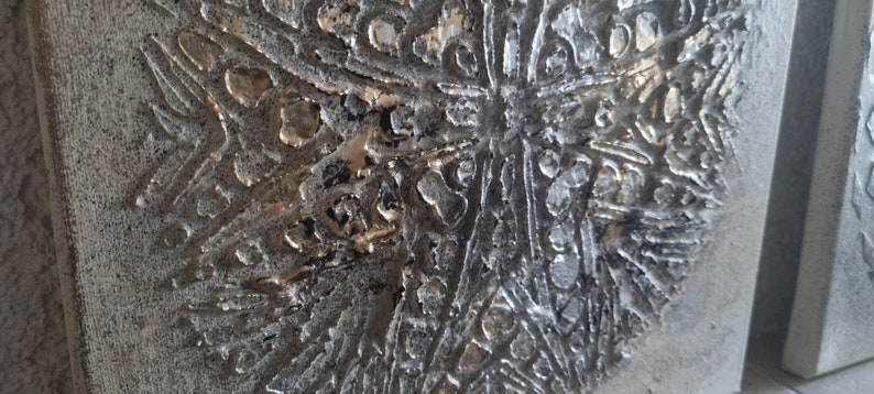 May include: A close-up of a textured, gray canvas with a raised, silver and gold design. The design is abstract and resembles a stylized flower or snowflake.