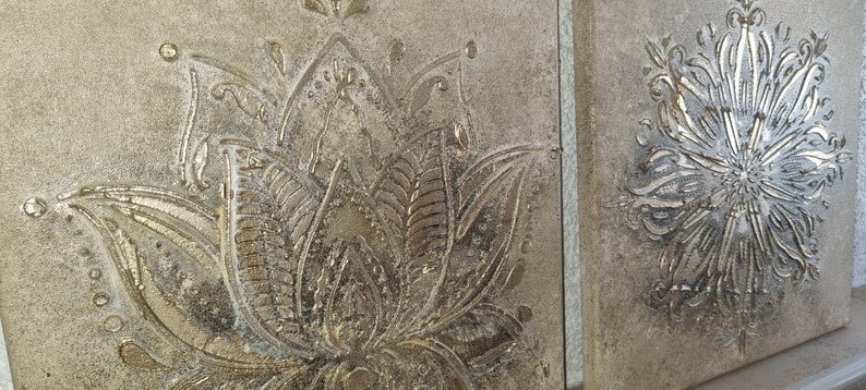 Op de afbeelding: Twee vierkante canvasdoeken met een reli&euml;fdesign. Het linker canvas heeft een zilverkleurig lotusbloemdesign. Het rechter canvas heeft een zilverkleurig bloemendesign.