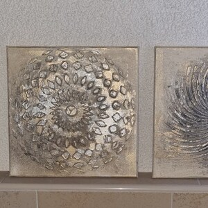 May include: Three square canvas wall art pieces with a silver and white abstract design. The designs are circular and have a textured look.
