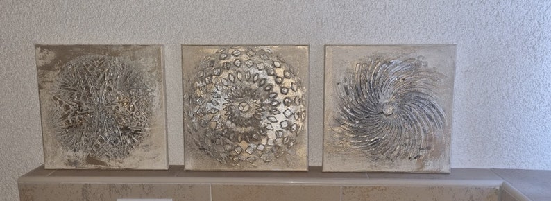 May include: Three square canvas wall art pieces with a silver and white abstract design. The designs are circular and have a textured look.
