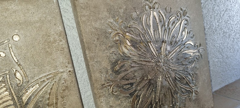 Op de afbeelding: Twee vierkante wanddecoraties van beige canvas met een reli&euml;f, gouden bloemmotief.