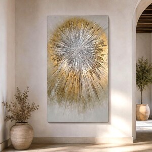 Pode incluir: Uma grande peça de arte de parede retangular com um design abstrato de raio de sol. O centro é composto por traços de tinta prateada e branca texturizada que irradiam para fora, rodeados por camadas de tinta dourada metálica sobre um fundo neutro.