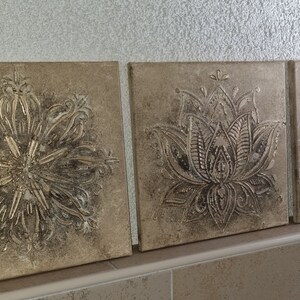 Op de afbeelding: Drie vierkante canvas wandkunststukken met een verweerde, beige achtergrond. Elk stuk heeft een zilveren metallic ontwerp van een mandala, een lotusbloem en een cirkelvormig patroon.
