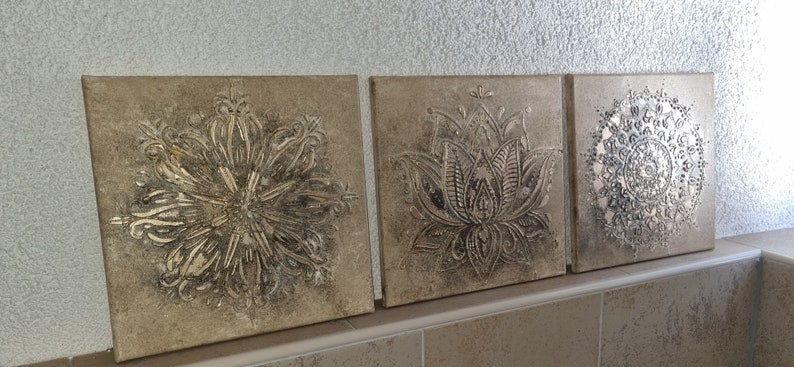 Op de afbeelding: Drie vierkante canvas wandkunststukken met een verweerde, beige achtergrond. Elk stuk heeft een zilveren metallic ontwerp van een mandala, een lotusbloem en een cirkelvormig patroon.