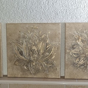 Op de afbeelding: Drie vierkante canvas wandkunststukken met een beige achtergrond en een verhoogd, reli&euml;fdesign in metallic goud. De ontwerpen zijn een mandala, een lotusbloem en een gestileerde bloem.