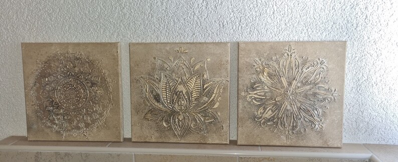 Op de afbeelding: Drie vierkante canvas wandkunststukken met een beige achtergrond en een verhoogd, reli&euml;fdesign in metallic goud. De ontwerpen zijn een mandala, een lotusbloem en een gestileerde bloem.