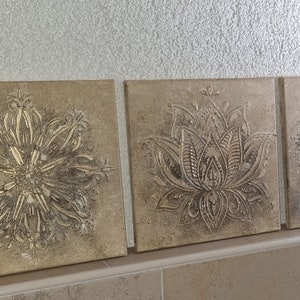Op de afbeelding: Drie vierkante canvas wandkunststukken met een beige achtergrond en zilveren metallic ontwerpen. De ontwerpen zijn ingewikkeld en omvatten een mandala, een lotusbloem en een geometrisch patroon.
