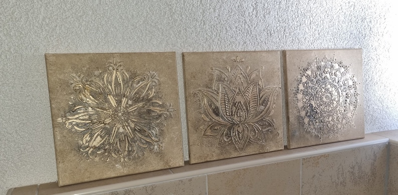 Op de afbeelding: Drie vierkante canvas wandkunststukken met een beige achtergrond en zilveren metallic ontwerpen. De ontwerpen zijn ingewikkeld en omvatten een mandala, een lotusbloem en een geometrisch patroon.