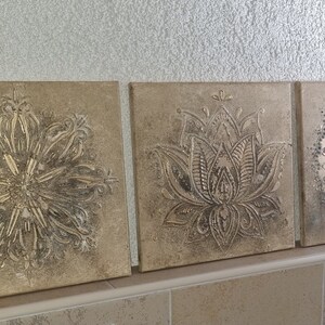 Op de afbeelding: Drie vierkante canvas wanddecoraties met een metallic gouden afwerking. De ontwerpen zijn ingewikkeld en bevatten een mandala, een lotusbloem en een geometrisch patroon.