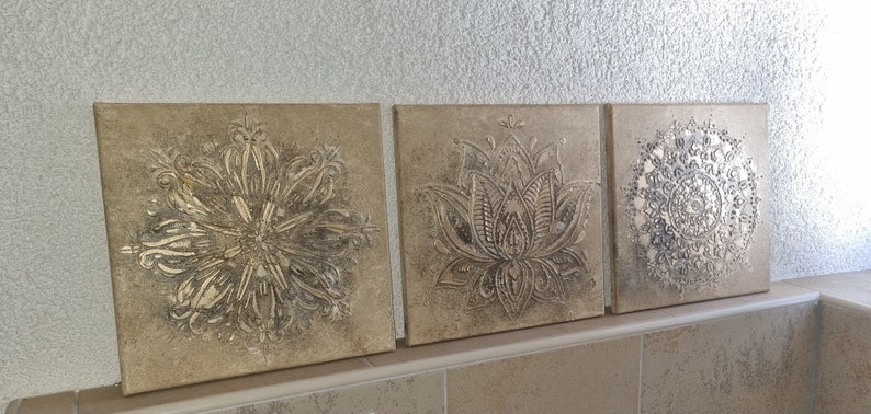 Op de afbeelding: Drie vierkante canvas wanddecoraties met een metallic gouden afwerking. De ontwerpen zijn ingewikkeld en bevatten een mandala, een lotusbloem en een geometrisch patroon.