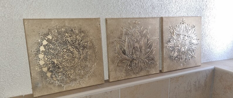 Op de afbeelding: Drie vierkante canvas wandkunststukken met een reli&euml;fdesign. Het ontwerp is zilverkleurig metallic op een beige achtergrond. De ontwerpen zijn een mandala, een lotusbloem en een gestileerde bloem.