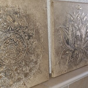 Op de afbeelding: Drie vierkante wanddecoraties met een verweerde, textuurrijke, beige achtergrond. Het middelste stuk heeft een zilveren reli&euml;f met een bloemenmotief. De twee zijstukken hebben een zilveren reli&euml;f met een mandala-ontwerp.