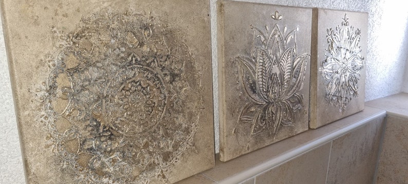 Op de afbeelding: Drie vierkante wanddecoraties met een verweerde, textuurrijke, beige achtergrond. Het middelste stuk heeft een zilveren reli&euml;f met een bloemenmotief. De twee zijstukken hebben een zilveren reli&euml;f met een mandala-ontwerp.