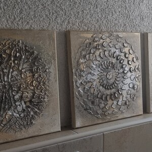 May include: Three square canvas paintings with a textured, metallic silver and gold finish. Each painting features a different circular design.
