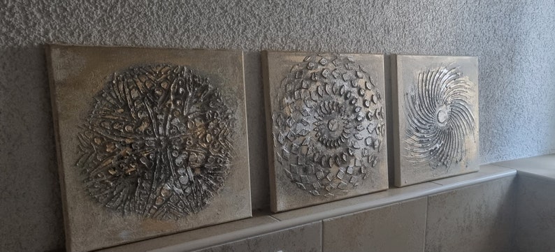 May include: Three square canvas paintings with a textured, metallic silver and gold finish. Each painting features a different circular design.