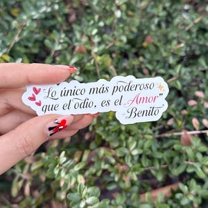May include: A white sticker with the quote "Lo único más poderoso que el odio, es el Amor" and the name "Benito" in black and pink script. The sticker has red heart accents and a star. The sticker is held in front of a green leafy background.