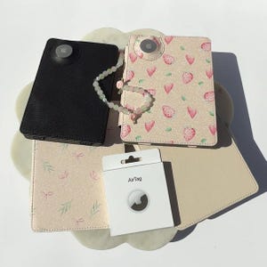 May include: A selection of AirTag cases in black, beige, and pink, with a matching bracelet. One case is black, another is patterned with strawberries and hearts, and a third has a bow and leaf design. A white AirTag box is also present.