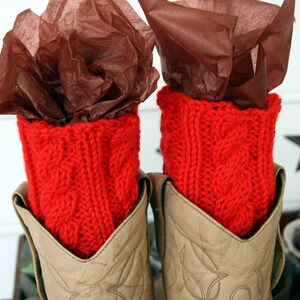 May include: Red knitted boot cuffs with a cable knit pattern, shown on a pair of brown cowboy boots.