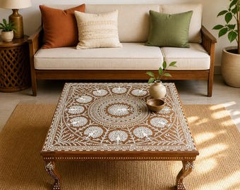 Pode incluir: Uma mesa de centro quadrada de madeira com um intrincado padrão incrustado em branco de motivos circulares e figuras de pavões estilizados na superfície superior, apoiada em quatro pernas curvas com detalhes florais brancos a condizer.