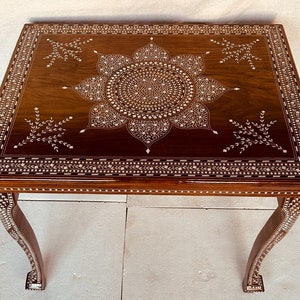 Op de afbeelding: Een bruine houten tafel met ingewikkelde bloemmotieven ingelegd met witte parelmoer. De tafel heeft vier poten met een gebogen ontwerp.
