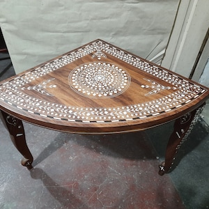 May include: A wooden corner table with intricate inlaid designs in white. The table has a rounded triangular top and three legs.